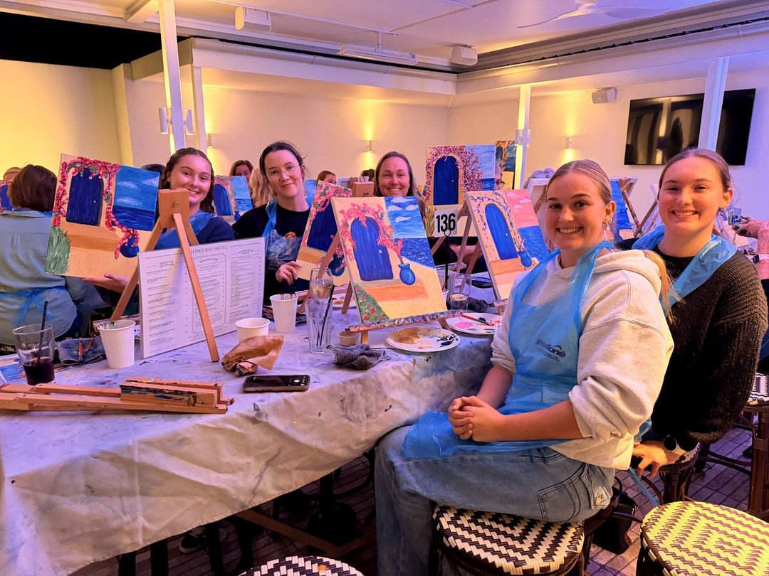 A group of Paint Juicy guests at their easels with finished Santorini blue door canvases at the 70s and 80s paint and sip night at The Shaws Bay Hotel Ballina