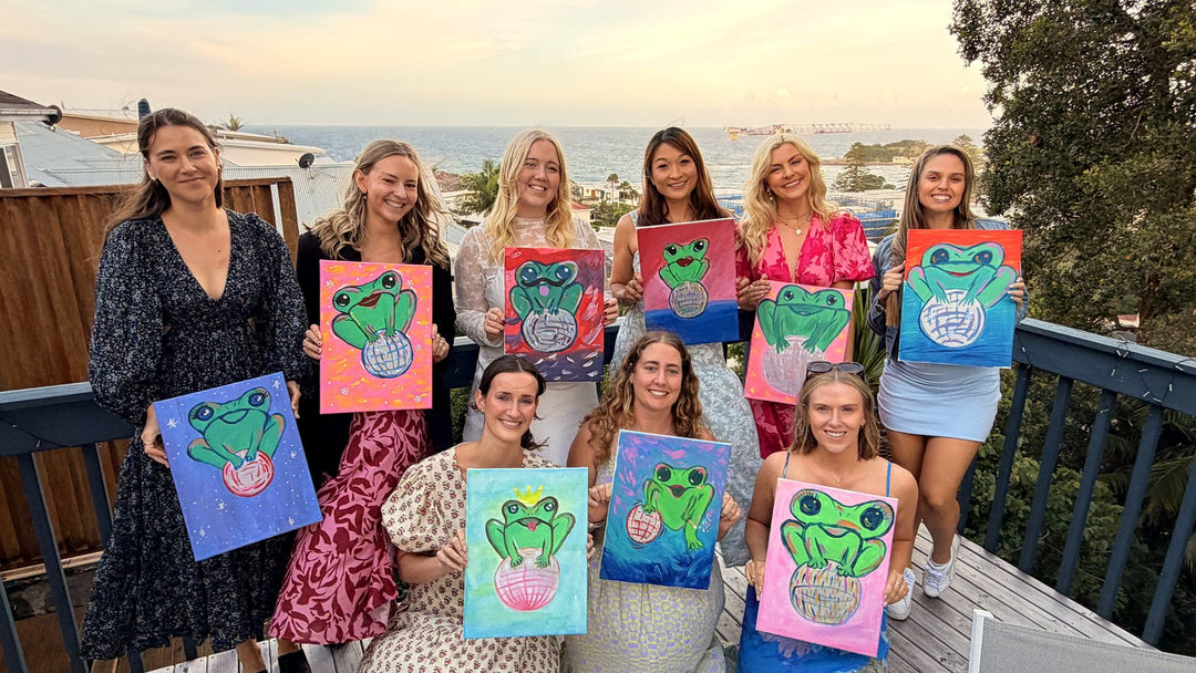 Ten women holding finished Disco Frog paintings on a Terrigal deck overlooking the ocean at a Paint Juicy private hens session