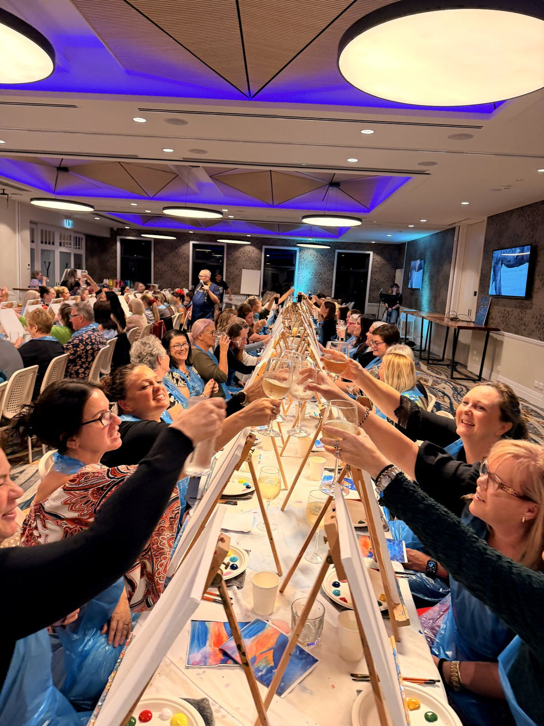 Paint and sip guests raising a toast along a long table at the Yarrabilba Hotel during a Paint Juicy 80s and 90s singalong night