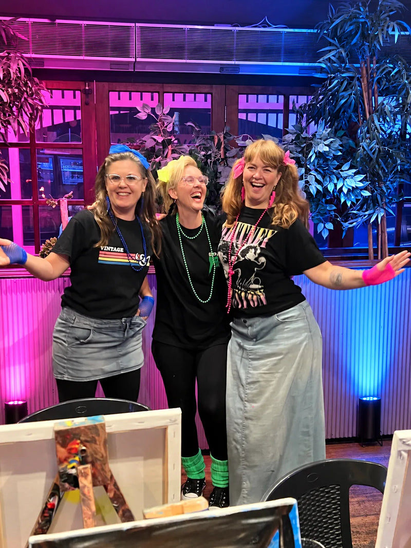 Three friends in 80s costume with neon accessories, laughing at an 80s-themed Paint Juicy paint and sip party