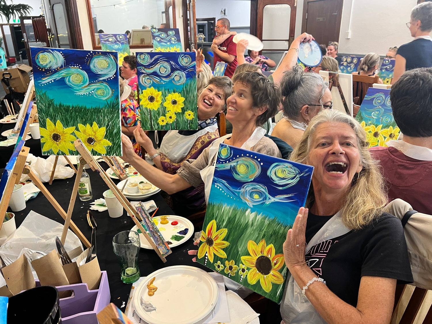 Group of people at a LISMORE painting class, holding up their artwork with sunflowers and starry night designs.