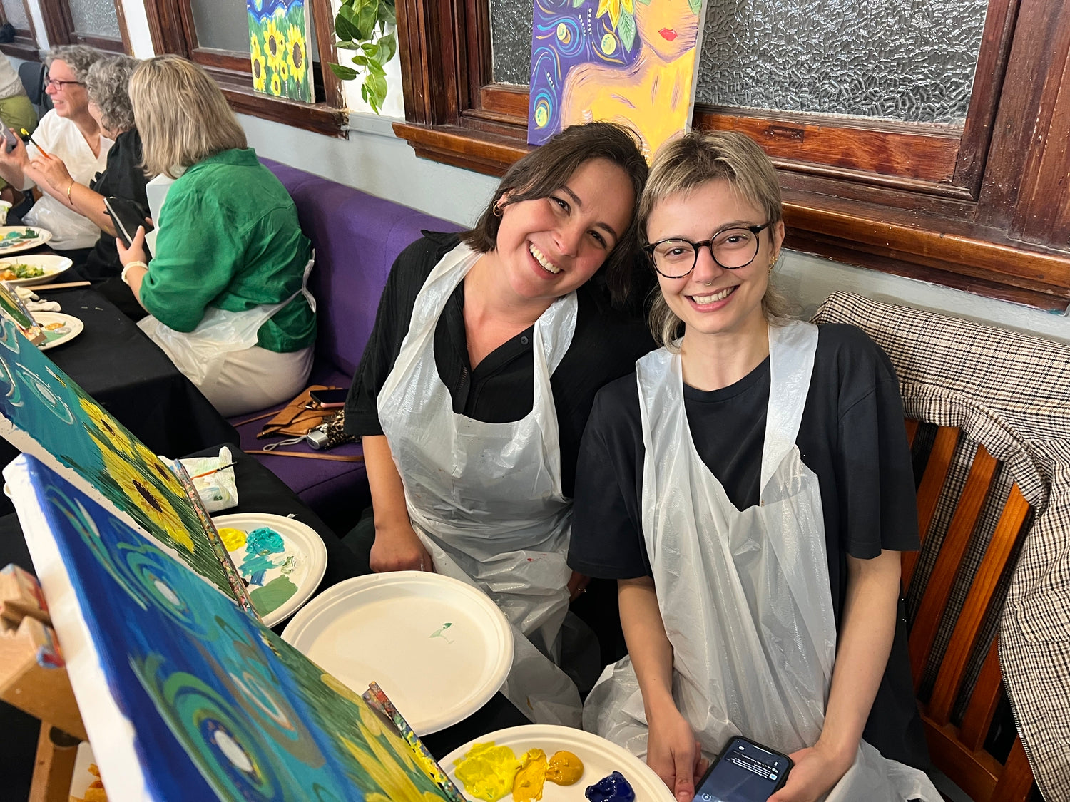 Two women in aprons sitting at a table with art supplies, smiling at the camera.