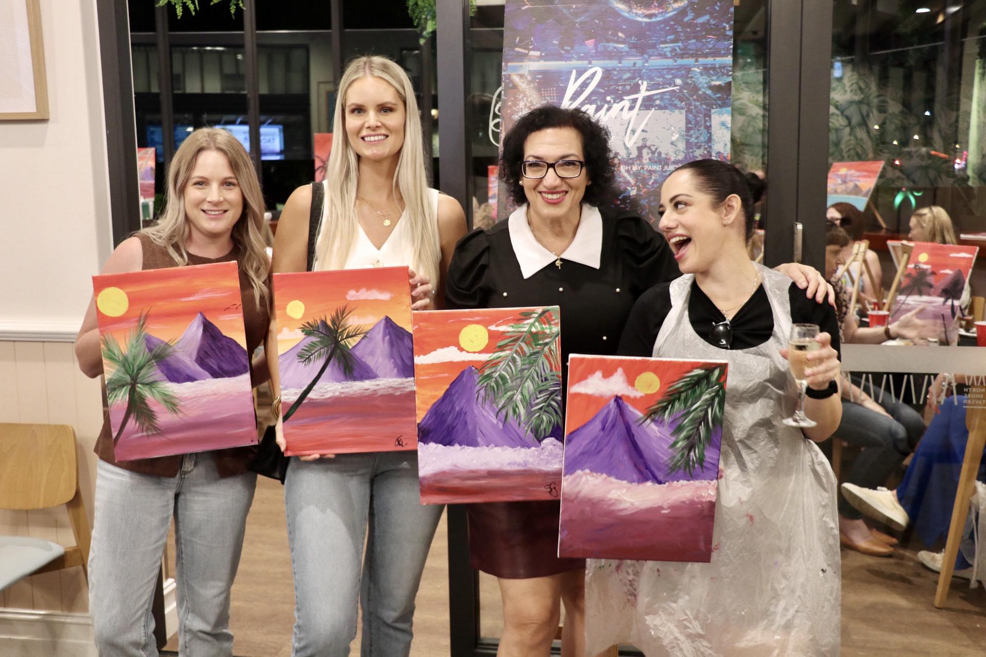 Four women holding up their painting of a sunset with palm trees, in an indoor setting at a Paint Juicy event.