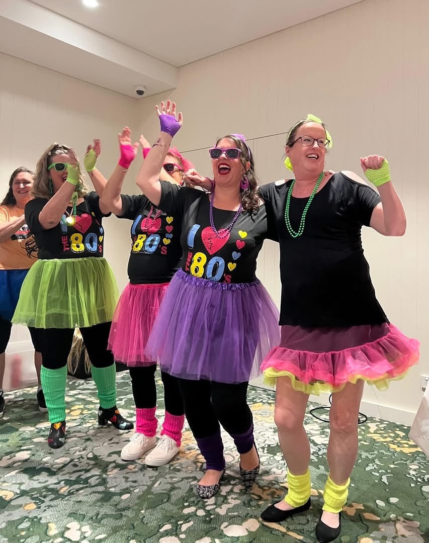 Group of people in colorful costumes and accessories, celebrating a 80s paint and sip theme.