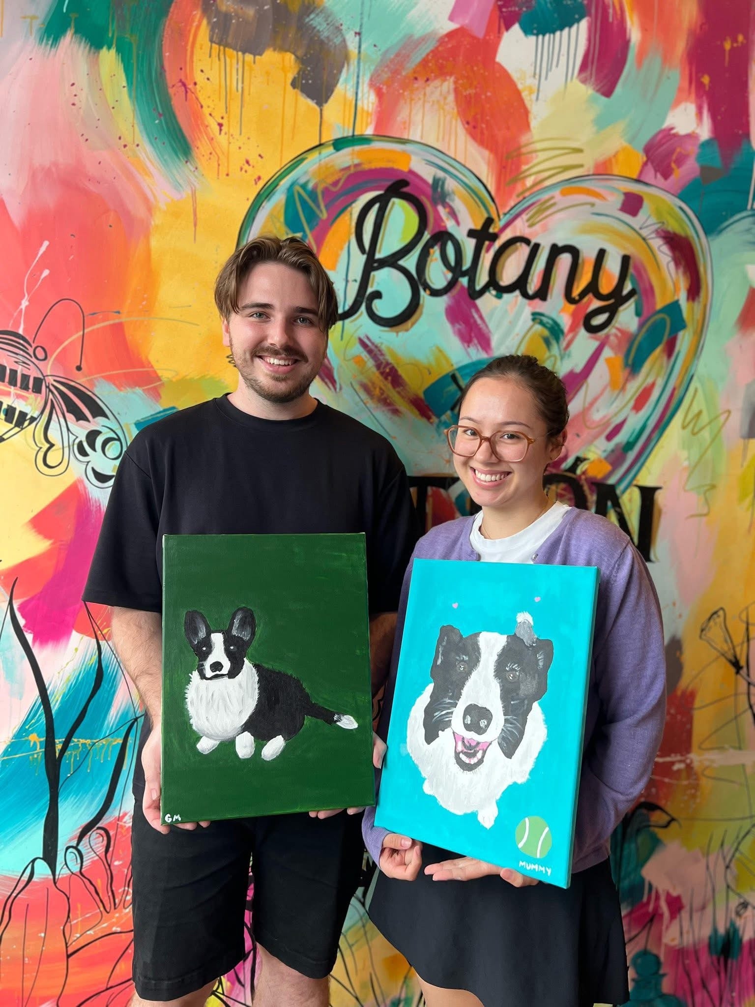 Two people holding paintings of dogs in front of a colourful mural 