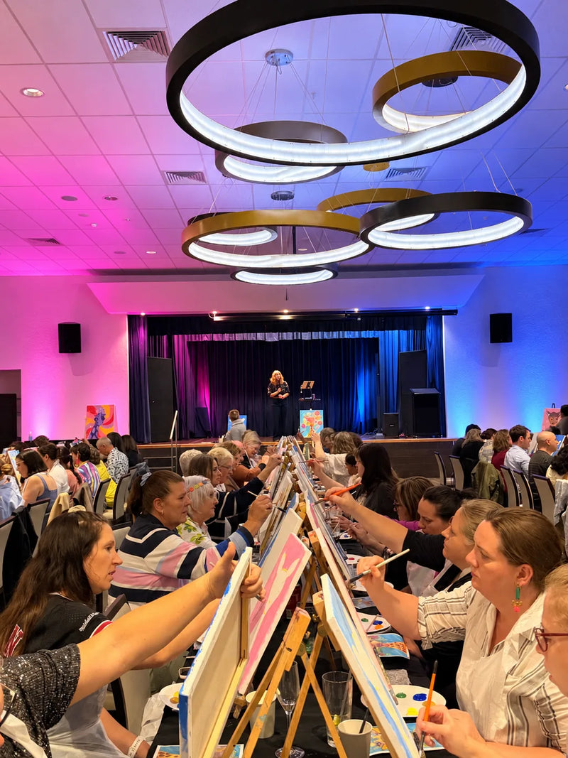 Guests painting at rows of easels under pink and blue lights at an Armidale paint and sip, host on stage