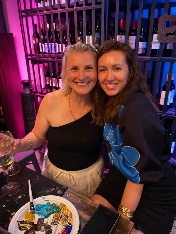 Two friends smiling beside glasses of white wine and a paint palette at a Paint Juicy paint and sip night out