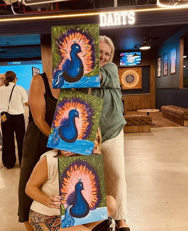 Three guests holding up their finished peacock paintings at a Paint Juicy session